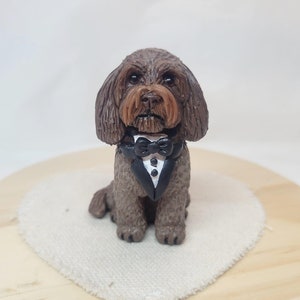 May include: A brown and black dog figurine wearing a black tuxedo with a white bow tie. The figurine is sitting on a white heart-shaped surface.