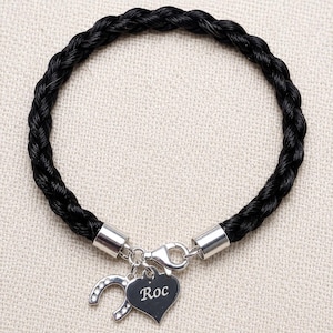 May include: A black braided bracelet featuring a silver-toned clasp and two dangling charms. One charm is a silver heart engraved with the name Roc, and the other charm is a silver horseshoe shape accented with small clear stones.