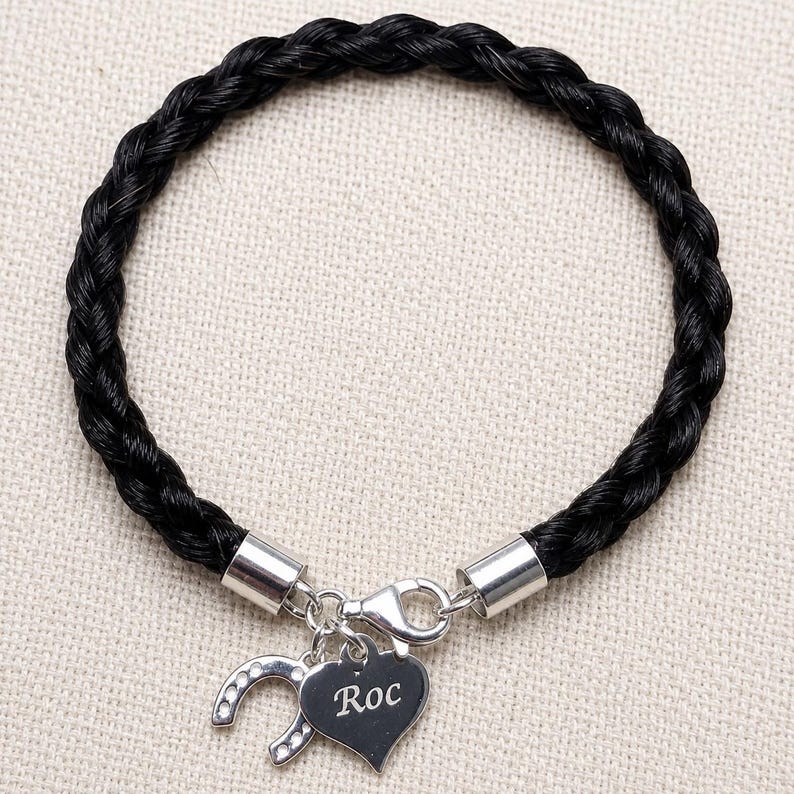 May include: A black braided bracelet featuring a silver-toned clasp and two dangling charms. One charm is a silver heart engraved with the name Roc, and the other charm is a silver horseshoe shape accented with small clear stones.