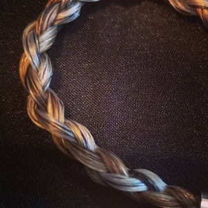 May include: A circular braided accessory featuring strands of metallic copper, silver, and dark grey fibers woven together in a tight, repeating pattern. The braid forms a continuous loop, showcasing a smooth, reflective texture throughout the design.