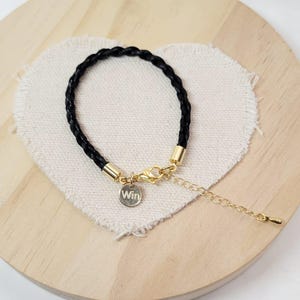 May include: Black braided leather bracelet with gold-coloured metal accents. A small, round charm with the word "Win" is attached. The bracelet is displayed on a heart-shaped fabric piece.