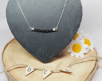 Puede incluir: Un collar de cordón de cuero negro con terminales de plata en una pantalla de pizarra en forma de corazón. El collar está sobre una base de madera con un borde de arpillera y una cadena de banderas de madera que deletrean "D.C.E."