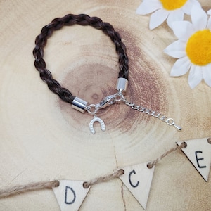 May include: A dark brown braided bracelet featuring silver-toned metal end caps, a lobster claw clasp, an adjustable chain, and a small silver-toned horseshoe charm. The bracelet is constructed from a braided material with a smooth, dark finish.