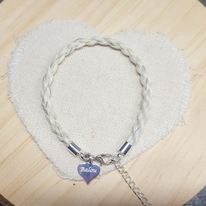 May include: A braided white hair bracelet featuring silver-toned metal end caps, a lobster claw clasp, and an adjustable chain. A small, heart-shaped silver-toned metal charm is attached to the clasp, engraved with the name Balou in white script.