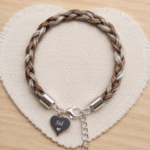 May include: A braided bracelet made of brown and white hair strands, featuring silver-toned metal end caps, a lobster claw clasp, and a heart-shaped charm engraved with the name Sid and a small heart symbol, attached to a short chain link.
