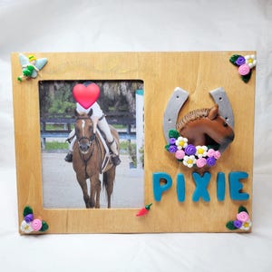 Può includere: Una cornice per foto in legno con una foto di una persona a cavallo. La cornice è decorata con un ferro di cavallo, una testa di cavallo, fiori e il nome "PIXIE" in lettere blu.