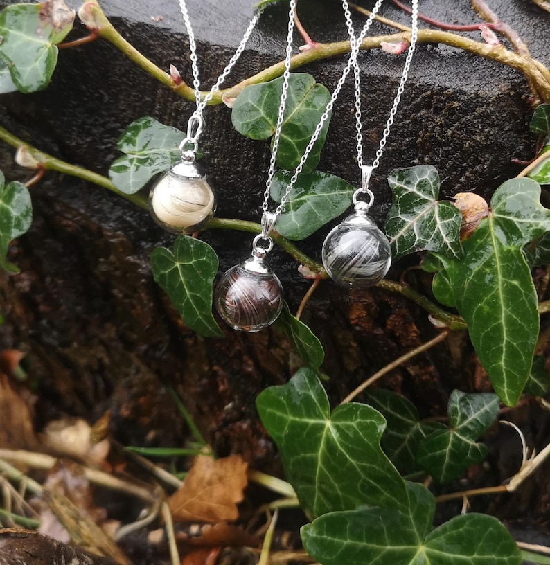 May include: Three silver necklaces with glass orbs containing strands of hair. The orbs are clear and the hair is visible inside. The necklaces are hanging from a tree branch.