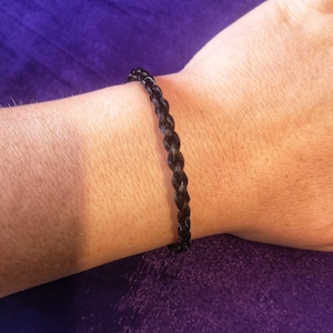 May include: A thin, dark brown bracelet featuring a braided texture. The bracelet is constructed from multiple strands woven together in a consistent, repeating pattern that encircles the wrist, creating a simple and uniform appearance.