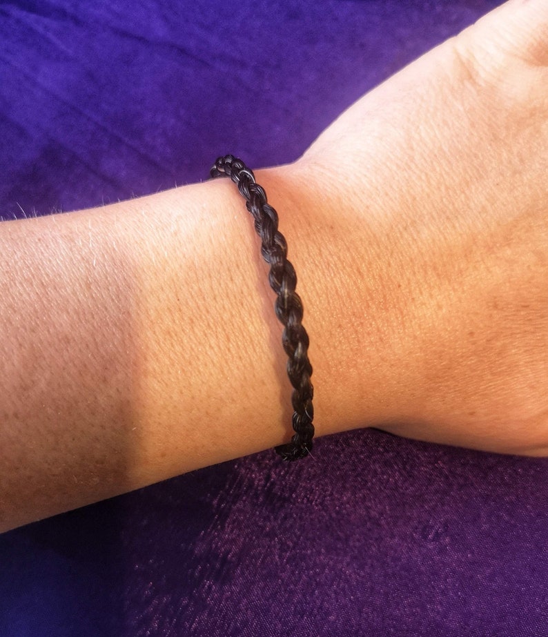 May include: A thin, dark brown bracelet featuring a braided texture. The bracelet is constructed from multiple strands woven together in a consistent, repeating pattern that encircles the wrist, creating a simple and uniform appearance.
