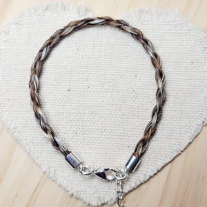 May include: A circular bracelet crafted from braided strands of brown, tan, and white hair. The ends of the braid are secured with polished silver-toned metal caps, featuring a silver-toned lobster claw clasp and a short adjustable chain link extension.