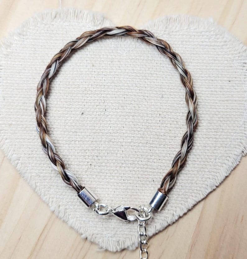 May include: A circular bracelet crafted from braided strands of brown, tan, and white hair. The ends of the braid are secured with polished silver-toned metal caps, featuring a silver-toned lobster claw clasp and a short adjustable chain link extension.
