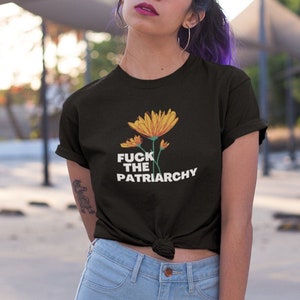 May include: Black t-shirt featuring a floral design with yellow and orange flowers and the text "FUCK THE PATRIARCHY" in white. The shirt is paired with light blue jeans.