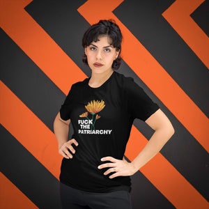 May include: Black t-shirt featuring a floral design with the text "FUCK THE PATRIARCHY" in white. The shirt has a crew neck and short sleeves. The background is a pattern of black and orange diagonal stripes.