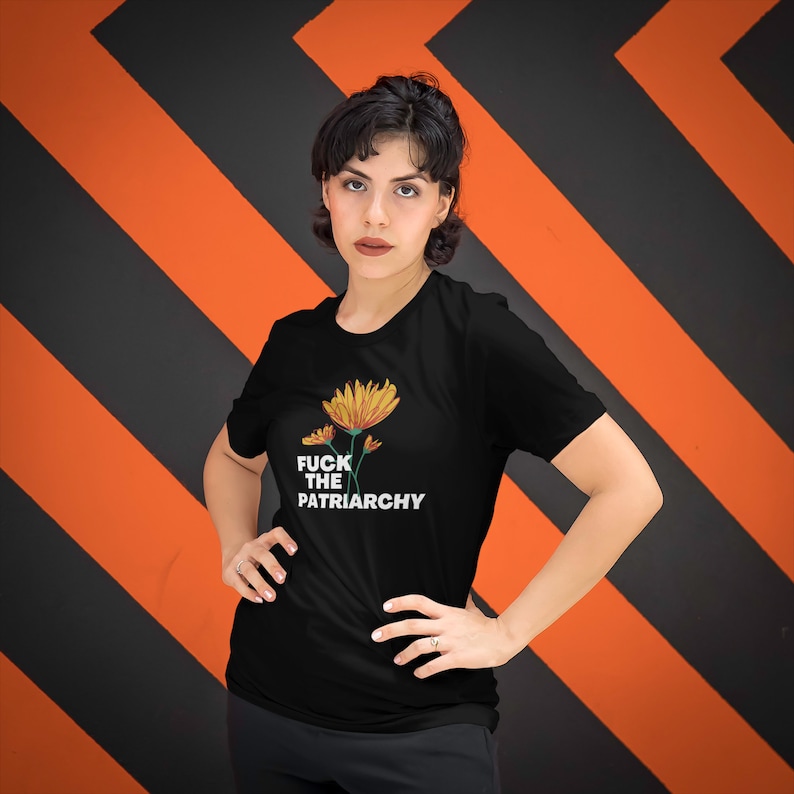 May include: Black t-shirt featuring a floral design with the text "FUCK THE PATRIARCHY" in white. The shirt has a crew neck and short sleeves. The background is a pattern of black and orange diagonal stripes.