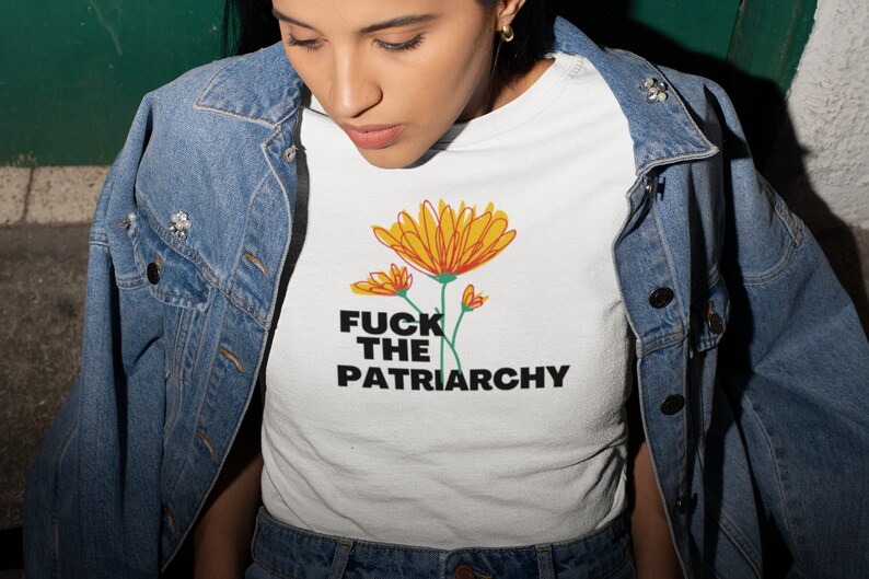 May include: White t-shirt with a floral design in yellow and orange, and the text "FUCK THE PATRIARCHY" in black. The shirt is worn with a denim jacket.