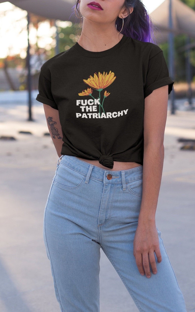May include: Black t-shirt featuring a floral design with yellow and orange flowers and the text "FUCK THE PATRIARCHY" in white. The shirt is paired with light blue jeans.