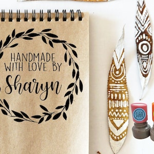 May include: A kraft paper notebook with a black wreath and the text "HANDMADE WITH LOVE BY Sharyn". Several stamps with ink pads and painted leaf art are also visible.