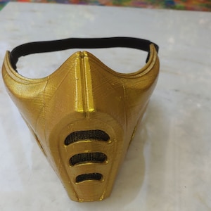 May include: A gold-colored plastic mask with a textured surface and a black strap. The mask has a design resembling a face with a mouth guard.