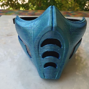 May include: A blue face mask with a textured surface and black mesh vents. The mask has a metallic sheen and is designed to cover the lower half of the face. The mask is likely a cosplay or costume accessory.