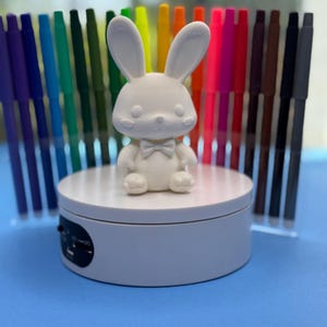 May include: A white, 3D-printed bunny figurine with a bow tie sits on a white rotating display stand. Behind the bunny are rows of colorful markers in various colors, including purple, green, yellow, orange, red, and brown. The background is a solid blue.