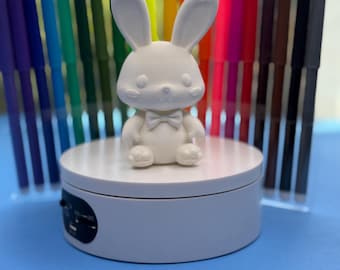 May include: A white, 3D-printed bunny figurine with a bow tie sits on a white rotating display stand. Behind the bunny are rows of colorful markers in various colors, including purple, green, yellow, orange, red, and brown. The background is a solid blue.