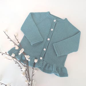 May include: A light blue knitted baby cardigan with white buttons and a ruffled hem.