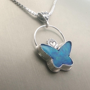 May include: A silver necklace with a butterfly pendant. The pendant features a blue butterfly design with a silver outline and a small heart detail. The necklace is displayed on a mannequin.