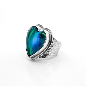 May include: A heart-shaped ring with a vibrant blue and green iridescent stone. The stone is set in a silver-toned, textured band. The ring's design features a hammered texture on the band, adding a unique touch to the jewelry.