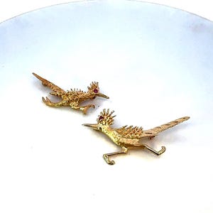 May include: Two gold-tone roadrunner pins with red rhinestone eyes. The roadrunners are in a running pose.