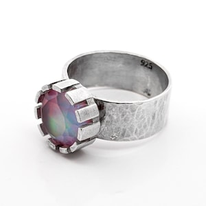 May include: A sterling silver ring with a large, iridescent gemstone. The ring features a wide band with a hammered texture and a bezel setting that holds the colorful stone. The ring is marked with "925".