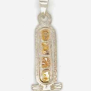 May include: A silver-toned pendant shaped like an Egyptian cartouche with a textured border. The center features four vertically stacked gold-toned hieroglyphic symbols. The pendant includes a top bail for attachment and a base with etched details.