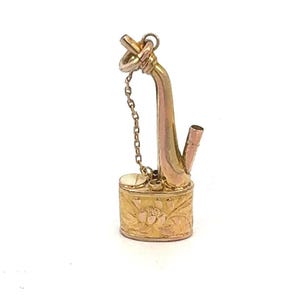 May include: A gold-toned pendant in the shape of a traditional Chinese pipe. The pipe is detailed with floral engravings and has a small chain attached to the top.