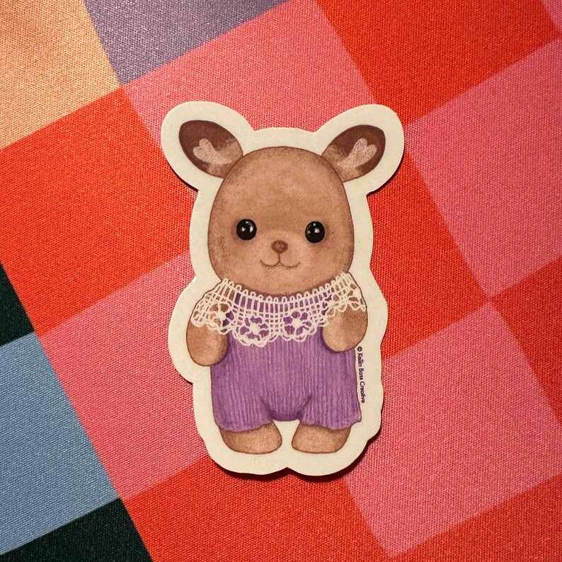 Op de afbeelding: Een gestanste sticker met een bruin antropomorf dierfiguur met grote zwarte ogen, gekleed in een witte kanten kraag en een paarse geribbelde romper. De tekst "Kalin Rose Crochet" is verticaal gedrukt aan de rechterkant van de sticker.