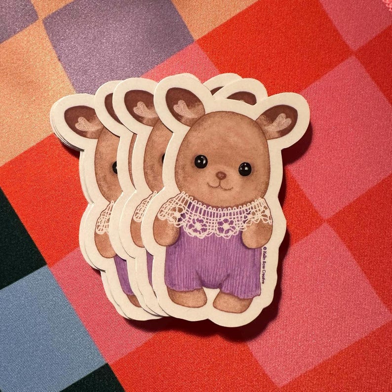 Op de afbeelding: Een stapel gestanste stickers met een bruin hert-personage met grote zwarte ogen, gekleed in een witte kanten kraag en een paarse tuinbroek. De illustratie bevat de tekst "Kalin Ross Creative" verticaal gedrukt aan de rechterkant.