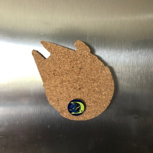 May include: A corkboard shaped like the Millennium Falcon from Star Wars with a blue and yellow enamel pin of a fish on it.