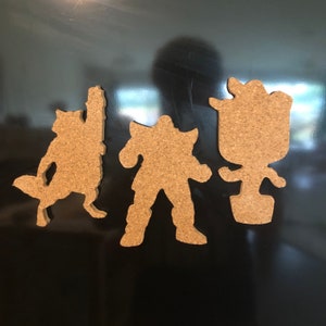 May include: Four corkboard silhouettes of characters, including a raccoon, a humanoid figure, and a plant. The silhouettes are brown and are attached to a black surface.