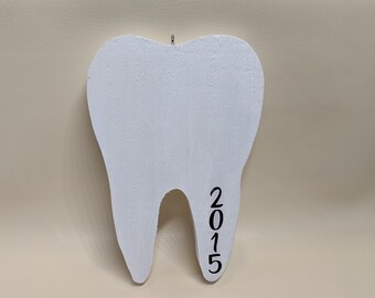 May include: A white, tooth-shaped ornament with the year "2015" in black script. The ornament has a small metal loop for hanging. The background is a solid, neutral color.