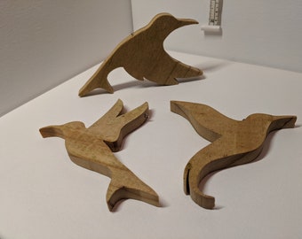 May include: Three wooden hummingbird decorations in various poses. The birds are cut from light-colored wood and have a natural finish. Each bird has a small metal loop for hanging. The image is taken against a white background.
