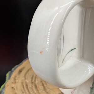 May include: Close-up of a white ceramic mug with a curved handle. The handle is smooth and glossy, with a few small imperfections. The mug appears to be sitting on a wooden surface.