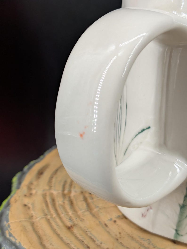 May include: Close-up of a white ceramic mug with a curved handle. The handle is smooth and glossy, with a few small imperfections. The mug appears to be sitting on a wooden surface.