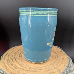 K&ouml;nnte beinhalten: Eine blaue Keramiktasse mit einem dekorativen Design. Die Tasse hat einen hellblauen Hintergrund mit einem gepunkteten Muster und einem Design aus Stern und Baum. Die Tasse steht auf einer Holzoberfl&auml;che.