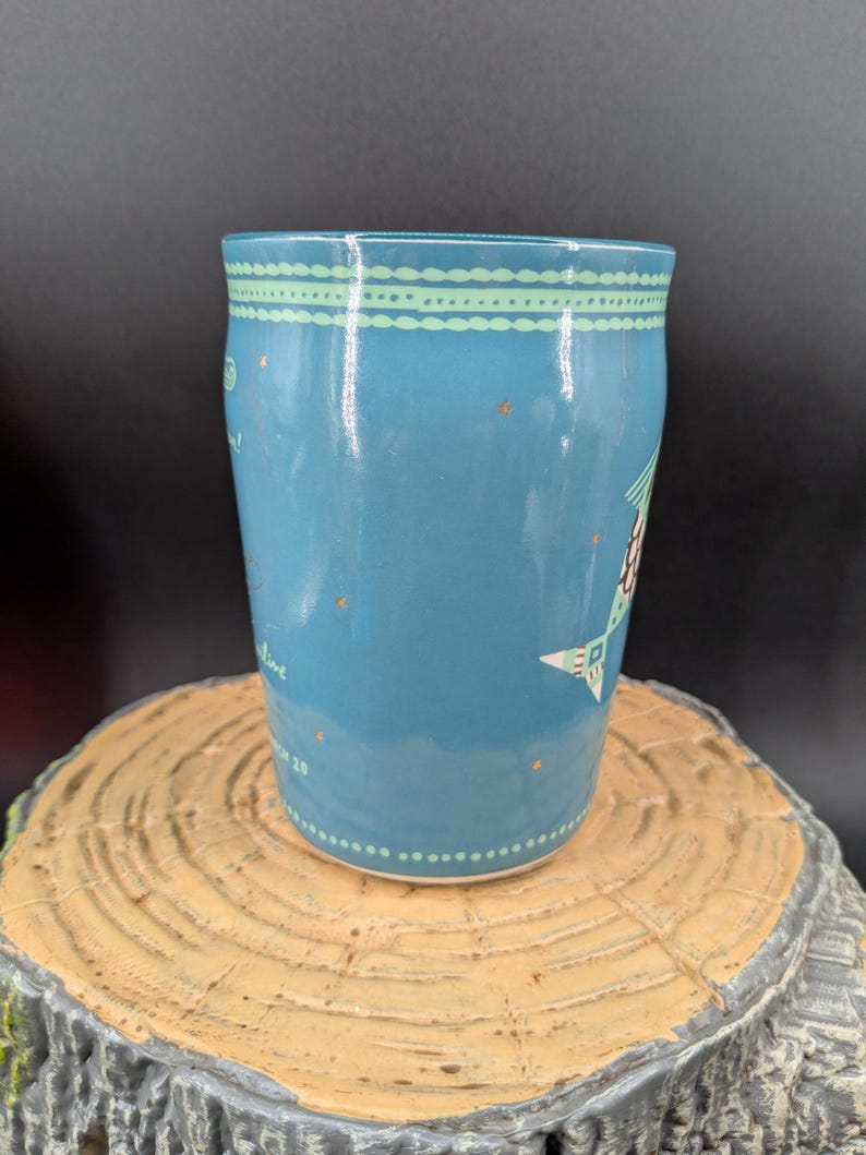 K&ouml;nnte beinhalten: Eine blaue Keramiktasse mit einem dekorativen Design. Die Tasse hat einen hellblauen Hintergrund mit einem gepunkteten Muster und einem Design aus Stern und Baum. Die Tasse steht auf einer Holzoberfl&auml;che.