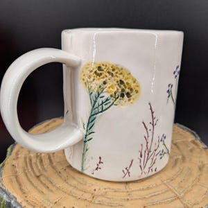 May include: White ceramic mug with a large, curved handle. The mug is decorated with a yellow and green floral design, along with smaller purple and red floral accents. The mug is sitting on a wooden surface.