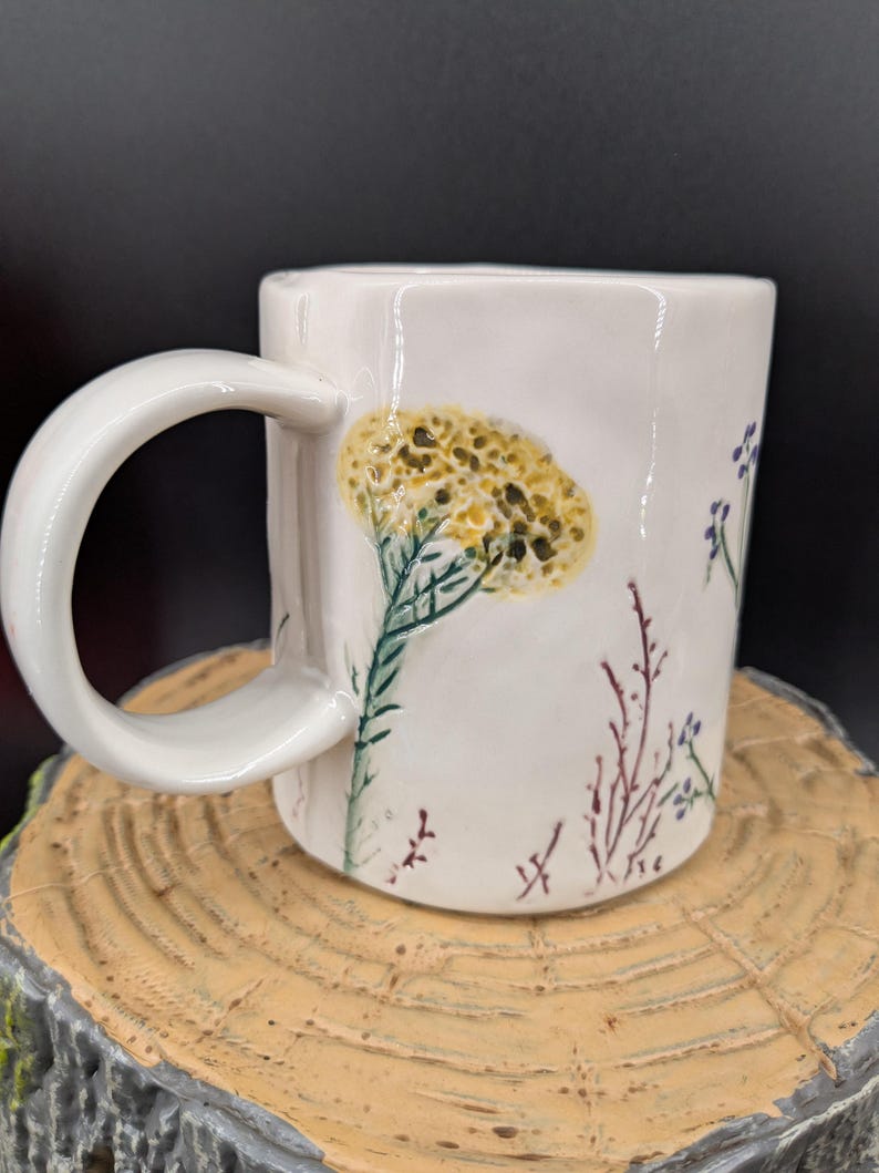 May include: White ceramic mug with a large, curved handle. The mug is decorated with a yellow and green floral design, along with smaller purple and red floral accents. The mug is sitting on a wooden surface.