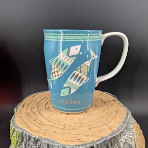 K&ouml;nnte beinhalten: Eine t&uuml;rkisfarbene Keramiktasse mit zwei stilisierten Fischen in Gold- und T&uuml;rkist&ouml;nen, die das Sternzeichen Fische darstellen. Die Tasse hat einen wei&szlig;en Henkel und ist mit Sternen und geometrischen Mustern verziert. Das Wort "PISCES" ist unten aufgedruckt.