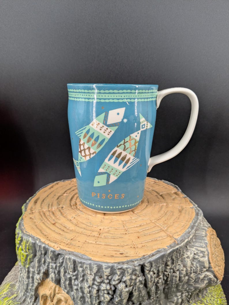 K&ouml;nnte beinhalten: Eine t&uuml;rkisfarbene Keramiktasse mit zwei stilisierten Fischen in Gold- und T&uuml;rkist&ouml;nen, die das Sternzeichen Fische darstellen. Die Tasse hat einen wei&szlig;en Henkel und ist mit Sternen und geometrischen Mustern verziert. Das Wort "PISCES" ist unten aufgedruckt.
