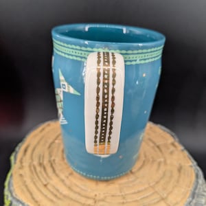 K&ouml;nnte beinhalten: Eine t&uuml;rkisfarbene Keramiktasse mit einem wei&szlig;en Griff, der mit goldenen Akzenten verziert ist. Die Tasse hat ein Stern- und geometrisches Muster in Wei&szlig; und Gr&uuml;n. Die Tasse steht auf einer Holzoberfl&auml;che.