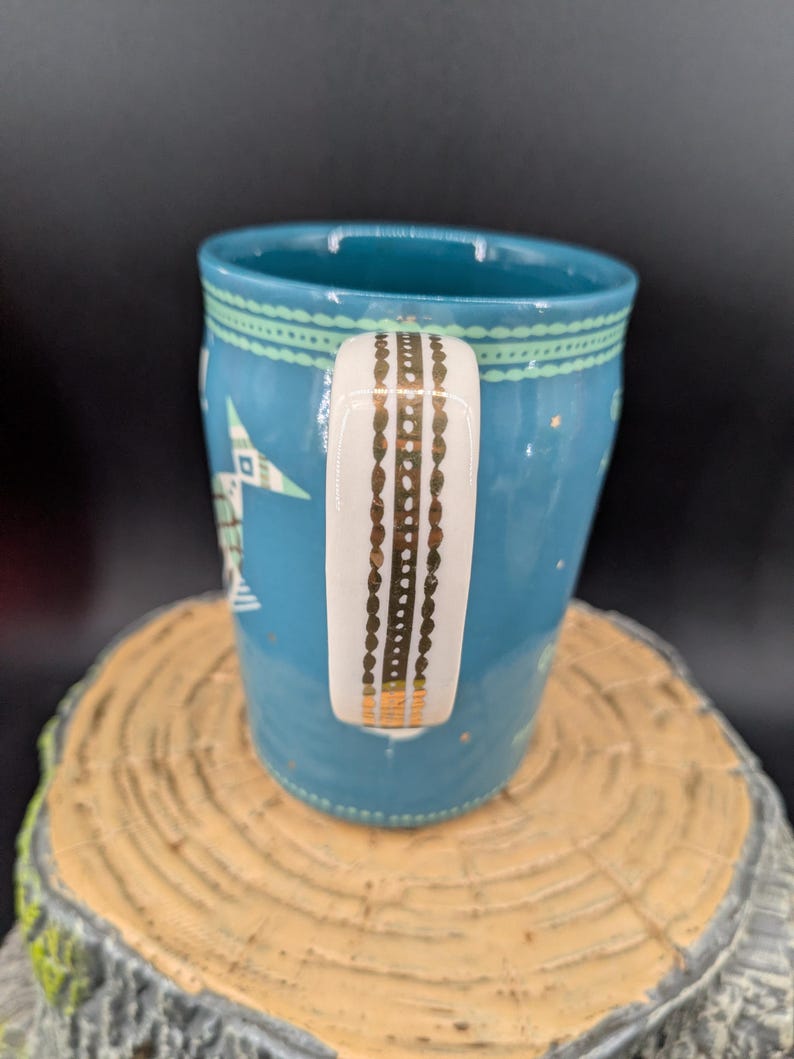K&ouml;nnte beinhalten: Eine t&uuml;rkisfarbene Keramiktasse mit einem wei&szlig;en Griff, der mit goldenen Akzenten verziert ist. Die Tasse hat ein Stern- und geometrisches Muster in Wei&szlig; und Gr&uuml;n. Die Tasse steht auf einer Holzoberfl&auml;che.