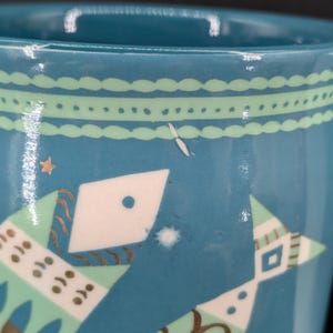 K&ouml;nnte beinhalten: Eine t&uuml;rkisfarbene Keramiktasse mit Henkel, verziert mit stilisierten Fisch- und Sternmustern in Wei&szlig;, Gr&uuml;n und Braun. Die Tasse hat ein hellgr&uuml;nes Band um den Rand.
