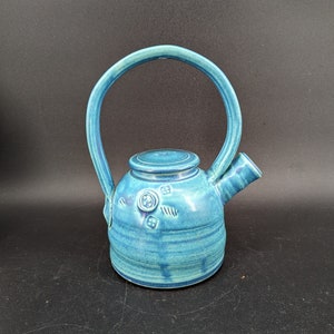 May include: A small, teal blue ceramic teapot with a rounded body and a curved handle. The teapot has a lid and a spout, and is decorated with a simple design of raised circles and lines.